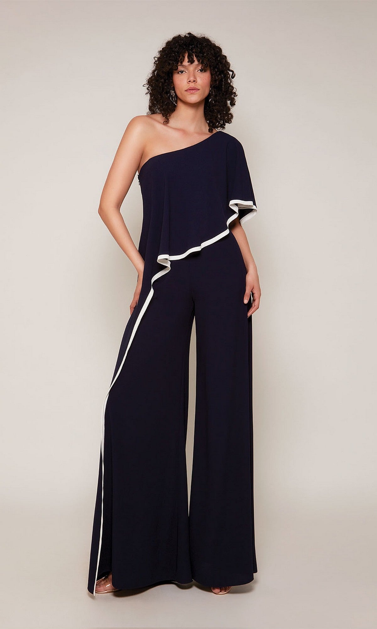 A woman stands facing forward, wearing the Long Formal Jumpsuit: Alyce 70049 in navy, featuring a one-shoulder design with wide legs and white trim outlining the diagonal neckline and leg seam.